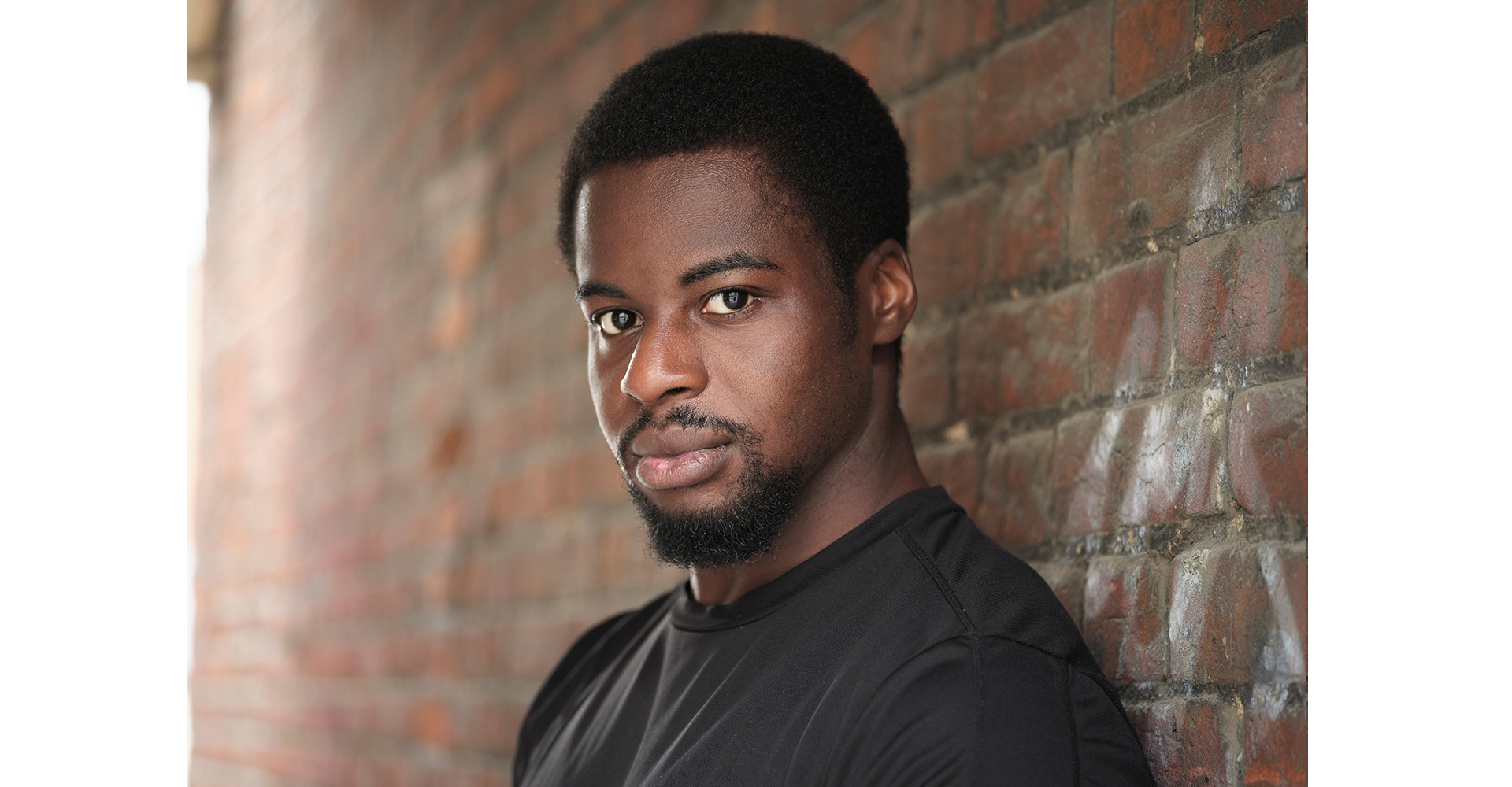 actor standing front brick wall in a black top actor head shot natural light